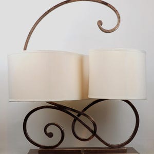 May include: A table lamp featuring two off-white cylindrical fabric shades supported by a dark bronze-toned metal base with intricate scrollwork. The base includes a tall, curved metal arm extending upwards and a rectangular metal foundation.