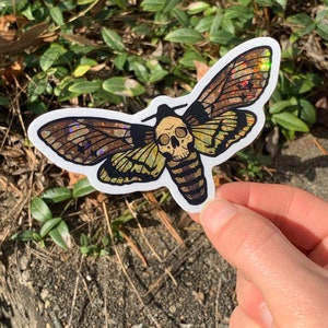 May include: A holographic sticker of a moth with a skull for a head. The moth has brown, green, and yellow wings with iridescent glitter.