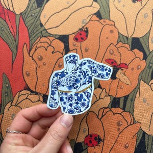 Puede incluir: Una pegatina con un diseño floral azul y blanco de una blusa con mangas largas y un cinturón dorado. La pegatina se sostiene frente a un fondo floral con tulipanes naranjas, hojas verdes y mariquitas.