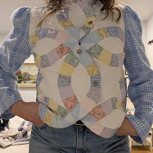 Peut inclure: Un gilet matelass&eacute; blanc avec un motif patchwork color&eacute;. Le gilet a un col montant et une fermeture &agrave; boutons. Le motif patchwork pr&eacute;sente une vari&eacute;t&eacute; de motifs floraux et g&eacute;om&eacute;triques dans des tons de rose, bleu, jaune et vert.