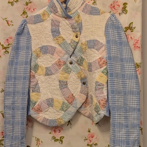 May include: A quilted jacket with a patchwork design in shades of pink, blue, yellow, and white. The jacket has a high collar and a button closure. The sleeves are made of a blue and white checked fabric.