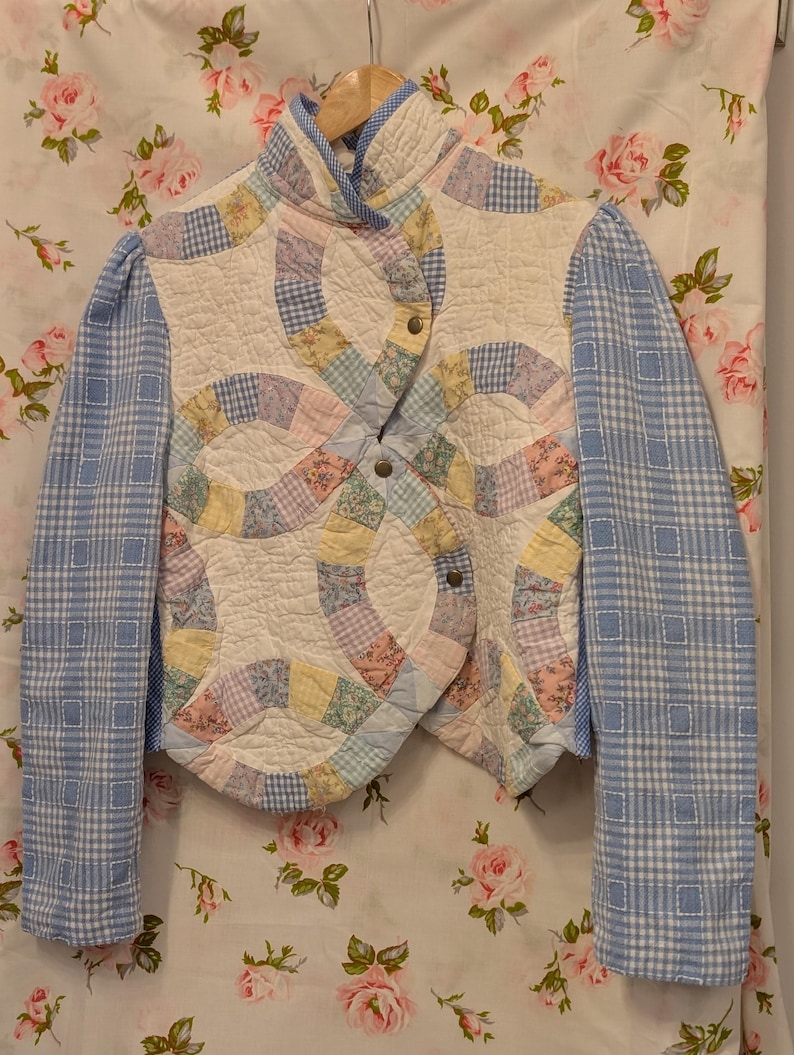 Peut inclure: Une veste matelass&eacute;e avec un motif patchwork dans des tons de rose, bleu, jaune et blanc. La veste a un col montant et une fermeture &agrave; boutons. Les manches sont en tissu &agrave; carreaux bleu et blanc.