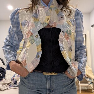 Peut inclure: Une veste en patchwork bleu et blanc avec un col montant et des manches bouffantes. La veste est en tissu matelass&eacute; et se ferme par des boutons. La veste est port&eacute;e sur un haut en maille noir et un jean bleu.