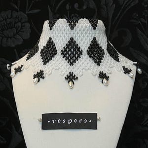 May include: A beaded choker necklace with a white and black geometric pattern, displayed on a white mannequin bust. The necklace has a scalloped edge with dangling pearls. A black tag with the word "vespers" is pinned to the bust.