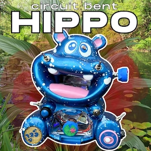 以下が含まれることがあります： 「circuit bent HIPPO」の白文字が入った、風変わりな青いカバの彫刻。カバはピンクの口、白い飛沫、さまざまな電子部品を持っています。背景には緑豊かな緑と池があります。