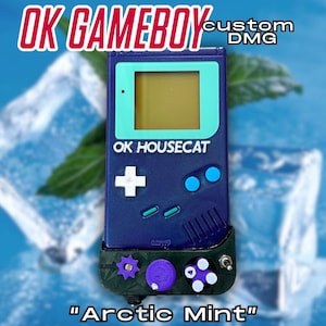 May include: A custom blue Game Boy DMG with a mint green screen frame and buttons. The text "OK GAMBOY" and "OK HOUSECAT" are visible. The bottom of the device has a custom green and purple design. The text "Arctic Mint" is at the bottom.