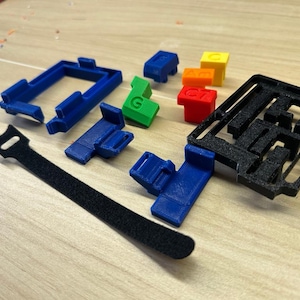 May include: An assortment of 3D-printed components in various colors, including blue, green, yellow, orange, and red. The items include a black Velcro strap, blue brackets, and a black puzzle-like structure. Some pieces have letters or numbers.