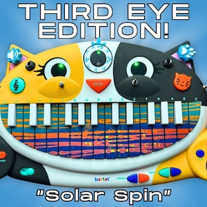 Op de afbeelding: Een katvormig elektronisch keyboard-speelgoed met de tekst THIRD EYE EDITION! en Solar Spin. Het apparaat heeft een geel en zwart gezicht, een mond van pianotoetsen met een blauw en oranje streeppatroon, een derde oog en een microfoon.