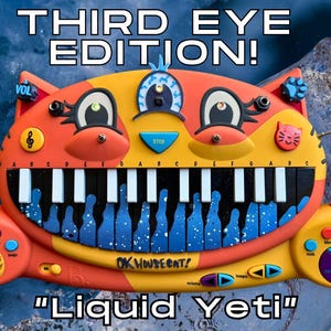 Puede incluir: Un juguete musical colorido con forma de gato, con teclado, botones y micrófono. El juguete es naranja, amarillo y azul, con el texto "THIRD EYE EDITION!" y "Liquid Yeti". El teclado tiene teclas blancas y detalles azules.