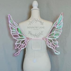 May include: A pair of iridescent pink and green butterfly wings with a white elastic band for attaching to the back. The wings are on a white dress form with the text "Minnow Lane Creations" on the front.