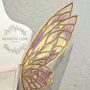 May include: A single iridescent pink and gold fairy wing with a detailed, intricate design. The wing is made of a lightweight material and has a delicate, ethereal look.