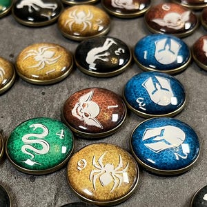May include: A collection of small, round, decorative game pieces with various designs. The pieces feature images of snakes, spiders, and other symbols in colors like green, gold, and blue. Each piece has a metallic edge.