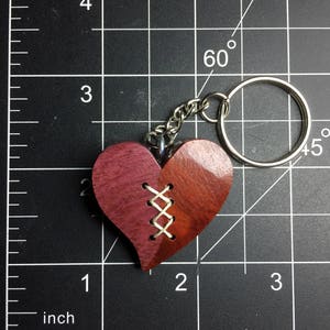May include: A wooden heart-shaped keychain with a stitched design, featuring a light brown and dark red wood grain pattern. The keychain is attached to a silver metal ring.