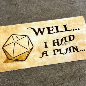 May include: A brown card with a drawing of a 20-sided die and the text "WELL... I HAD A PLAN..."