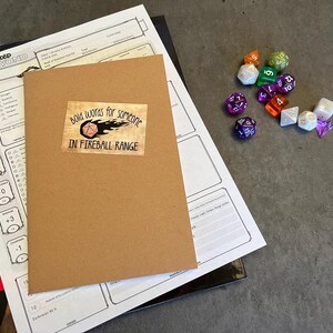 May include: A brown notebook with a sticker that says "Bold words for someone in fireball range". The sticker features a cartoon image of a fireball and a 20-sided die. The notebook is on top of a white sheet of paper with a grid pattern.