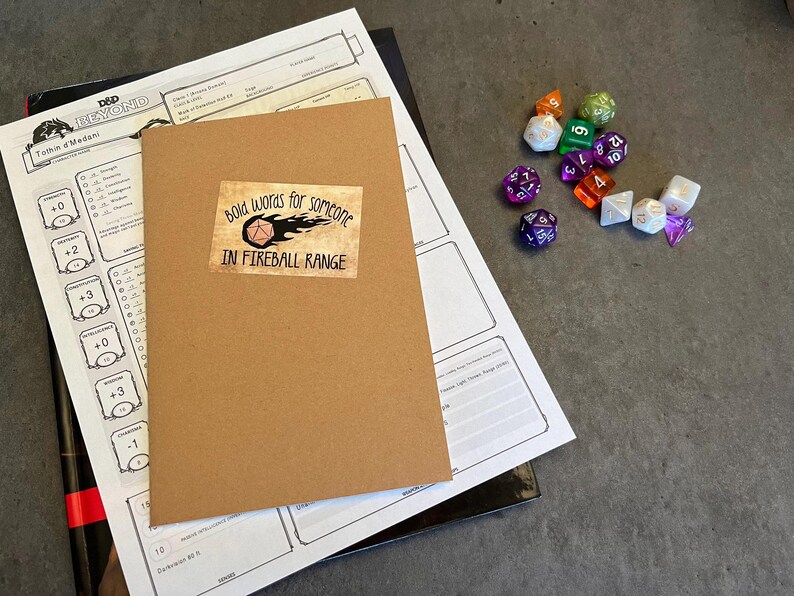 May include: A brown notebook with a sticker that says "Bold words for someone in fireball range". The sticker features a cartoon image of a fireball and a 20-sided die. The notebook is on top of a white sheet of paper with a grid pattern.