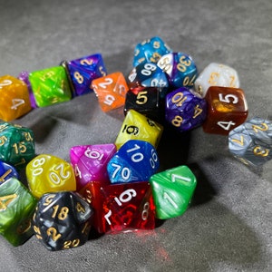 May include: A collection of colourful, iridescent dice with gold numbers. The dice are arranged in a circle on a grey surface.