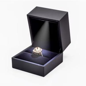 May include: A black, square ring box with a hinged lid, open to reveal a diamond ring on a dark blue velvet cushion. The ring is illuminated by a spotlight from the top of the box, highlighting its intricate design and sparkling facets.