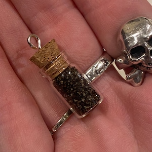 Op de afbeelding: Een klein glazen flesje met een kurkstop, gevuld met zwart zand, hangt aan een zilveren ketting. Het flesje bevindt zich op een hand die een zilveren ring met een schedelontwerp draagt.