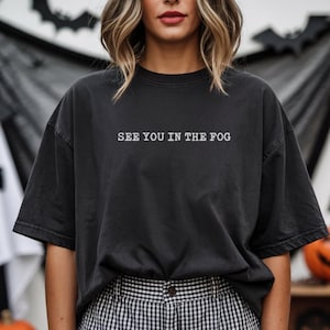 May include: A charcoal black short-sleeve t-shirt featuring the white text "SEE YOU IN THE FOG" printed across the chest. The garment has a crew neckline, a relaxed fit, and a slightly faded, washed-out appearance throughout the fabric.