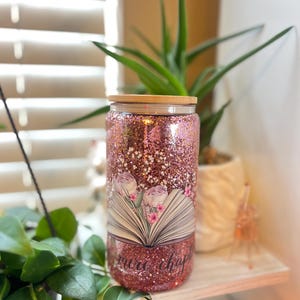 May include: A pink glitter tumbler with a wood lid and a design of an open book with flowers and the text "more chapters".
