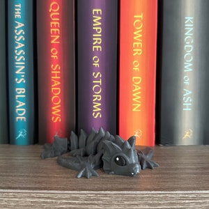 May include: A black, 3D printed dragon figurine with a spiky back and a toothy grin. The dragon is sitting on a wooden shelf in front of a row of books with the titles "Kingdom of Ash", "Tower of Dawn", "Empire of Storms", "Queen of Shadows", and "The Assassin's Blade".