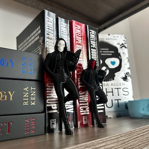 Könnte beinhalten: Zwei schwarze Figuren einer Person in Kapuzenkleidung, eine mit weißer Totenkopfmaske und die andere mit roter Maske, stehen vor einer Reihe von Büchern. Die Bücher tragen Titel wie "Penelope Douglas" und "Lights Out."