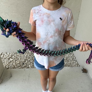 Puede incluir: Un niño sostiene un largo dragón de origami iridiscente hecho de papel doblado. El dragón es verde, morado y azul con un degradado de color. El dragón mide aproximadamente 90 cm de largo.