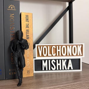 Puede incluir: Una figura negra de una persona con chaqueta y pantalones est&aacute; junto a libros. Los libros se titulan "Monster" y "Hunt the Villain". Tambi&eacute;n hay un letrero con las palabras "Volchonok" y "Mishka".