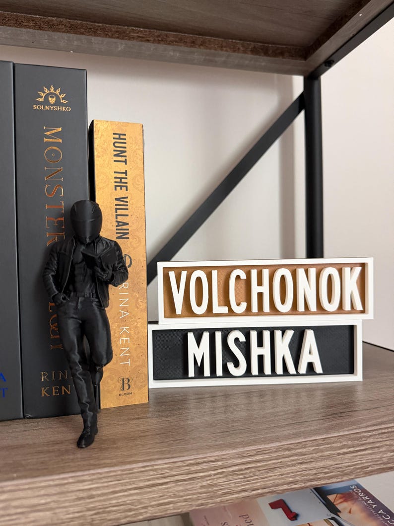 Puede incluir: Una figura negra de una persona con chaqueta y pantalones est&aacute; junto a libros. Los libros se titulan "Monster" y "Hunt the Villain". Tambi&eacute;n hay un letrero con las palabras "Volchonok" y "Mishka".