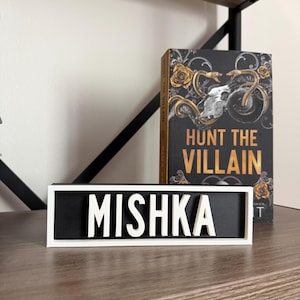 Puede incluir: Un letrero blanco y negro con el nombre "MISHKA" en letras blancas y en negrita. El letrero est&aacute; sobre una estanter&iacute;a de madera, junto a un libro titulado "HUNT THE VILLAIN" con una cubierta negra y detalles dorados.