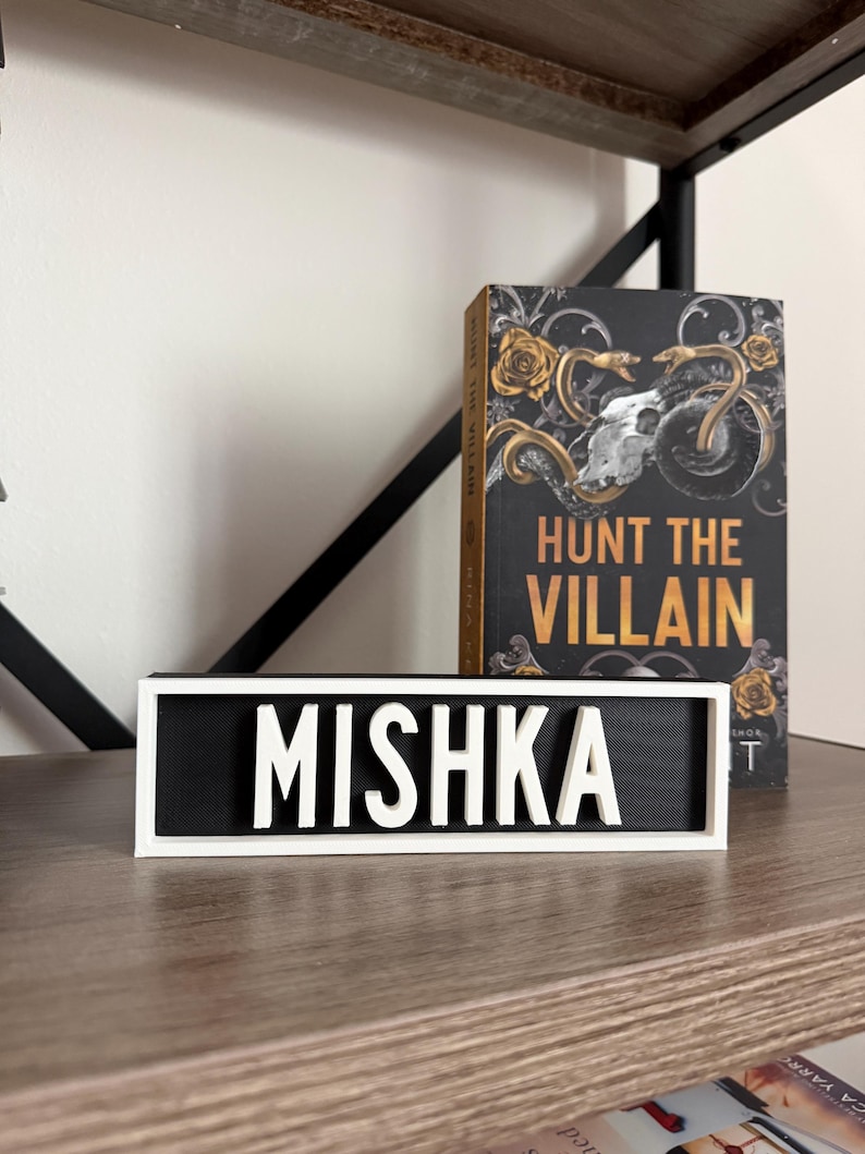 Puede incluir: Un letrero blanco y negro con el nombre "MISHKA" en letras blancas y en negrita. El letrero est&aacute; sobre una estanter&iacute;a de madera, junto a un libro titulado "HUNT THE VILLAIN" con una cubierta negra y detalles dorados.