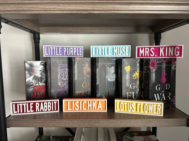 Może przedstawiać: P&oacute;łka prezentuje książki i dekoracyjne znaki. Znaki wyświetlają teksty "LITTLE PURPLE", "LITTLE MUSE", "MRS. KING", "LITTLE RABBIT", "LISICHKA" i "LOTUS FLOWER". Książki mają tytuły takie jak "GOD OF WAR" i "GOD OF FUR".
