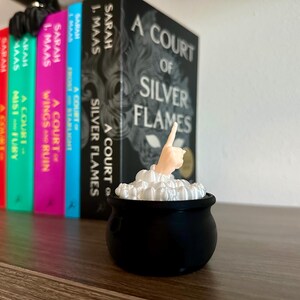 Op de afbeelding: Een zwarte ketel met wit schuim en een hand die uit het schuim steekt, wijzend naar boven. De ketel staat voor een boekenkast met een reeks boeken getiteld "A Court of..." door Sarah J. Maas.