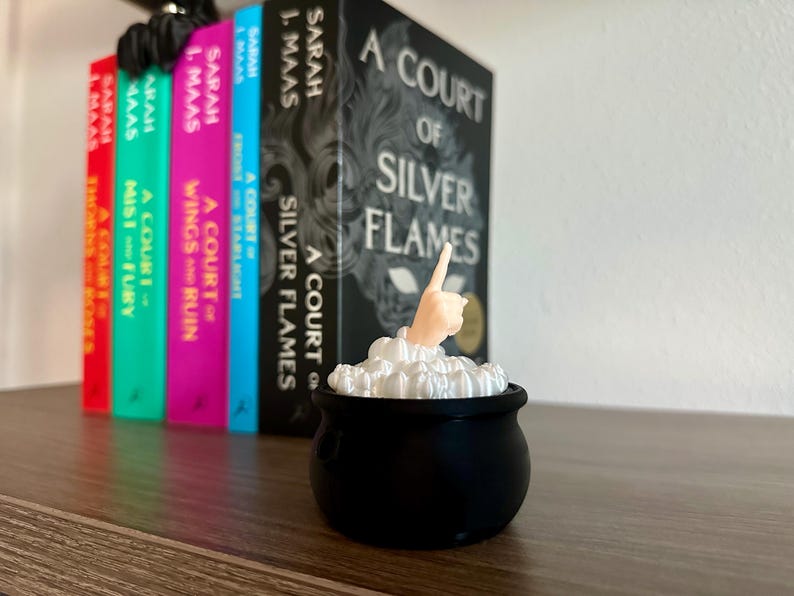 Op de afbeelding: Een zwarte ketel met wit schuim en een hand die uit het schuim steekt, wijzend naar boven. De ketel staat voor een boekenkast met een reeks boeken getiteld "A Court of..." door Sarah J. Maas.