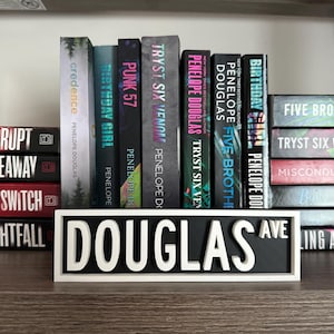 Puede incluir: Un letrero blanco con letras negras que dice "DOUGLAS AVE" se muestra en un estante con una colección de libros. Los libros son principalmente negros con letras blancas y tienen títulos como "Birthday Girl", "Five Brothers", "Tryst Six Venom" y "Misconduct".