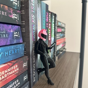 May include: A black figurine of a person in a motorcycle helmet and outfit, leaning against a shelf of books. The books have titles like "TRYST SIX VENOM" and "PUNK 57". The figurine is holding a small device.