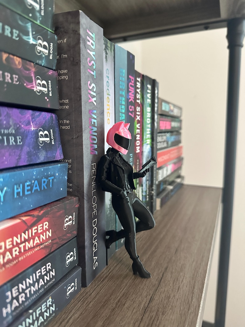 May include: A black figurine of a person in a motorcycle helmet and outfit, leaning against a shelf of books. The books have titles like "TRYST SIX VENOM" and "PUNK 57". The figurine is holding a small device.