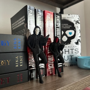 Puede incluir: Dos figuras negras de figuras encapuchadas con m&aacute;scaras de calavera, apoyadas en una estanter&iacute;a de libros. Una tiene una m&aacute;scara blanca, la otra una m&aacute;scara roja. Los libros incluyen t&iacute;tulos como "Corrupt" y "Lights Out".