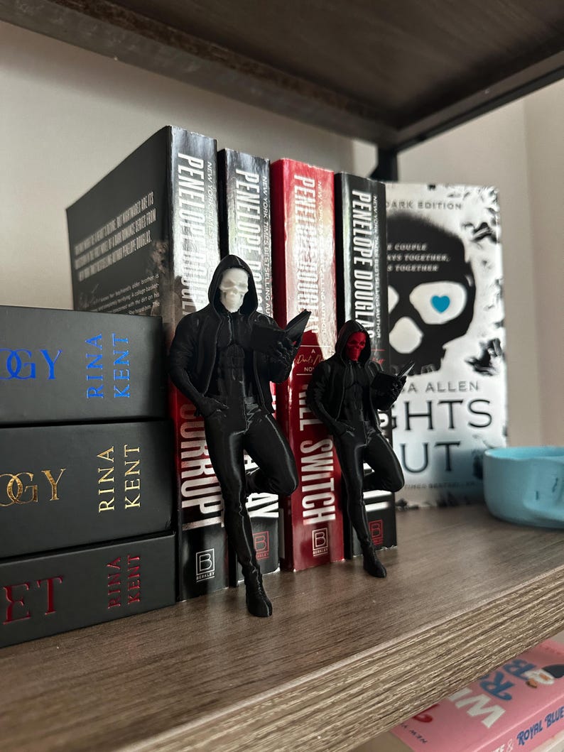 Puede incluir: Dos figuras negras de figuras encapuchadas con m&aacute;scaras de calavera, apoyadas en una estanter&iacute;a de libros. Una tiene una m&aacute;scara blanca, la otra una m&aacute;scara roja. Los libros incluyen t&iacute;tulos como "Corrupt" y "Lights Out".