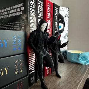 Puede incluir: Soportes para libros negros con forma de calavera, con forma de persona, apoyados en una fila de libros. Los libros tienen t&iacute;tulos de Penelope Douglas y Rina Kent. Tambi&eacute;n se ve un portavelas azul con forma de calavera.