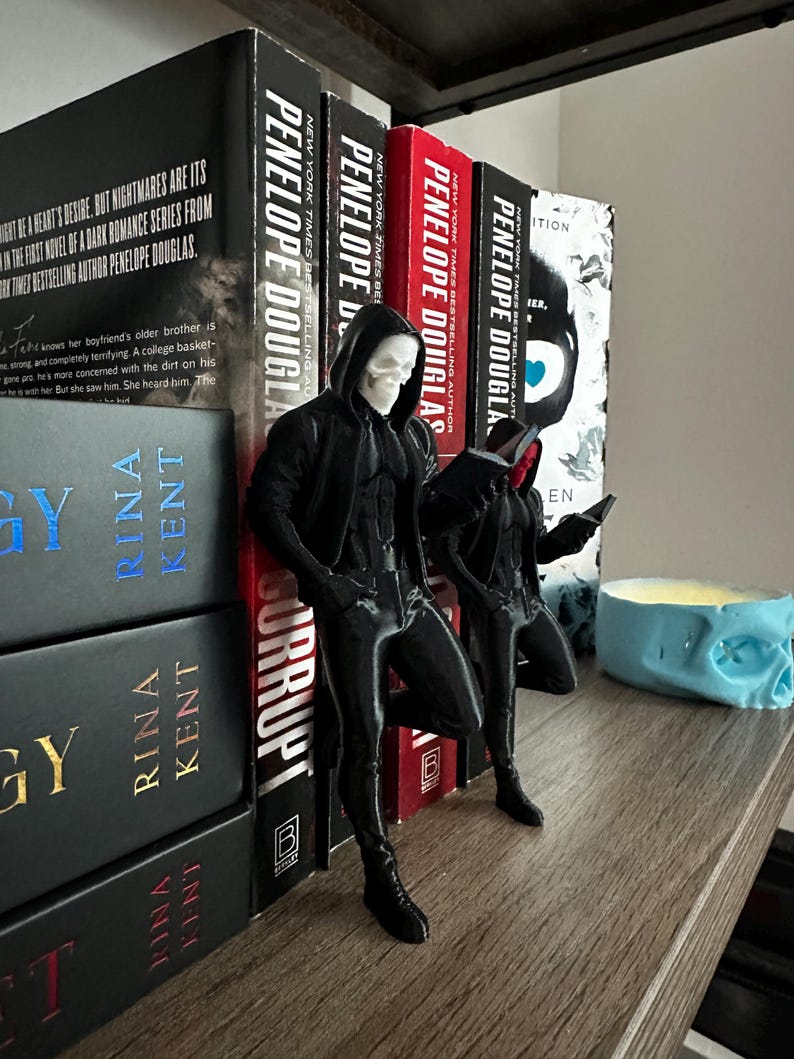 Puede incluir: Soportes para libros negros con forma de calavera, con forma de persona, apoyados en una fila de libros. Los libros tienen t&iacute;tulos de Penelope Douglas y Rina Kent. Tambi&eacute;n se ve un portavelas azul con forma de calavera.