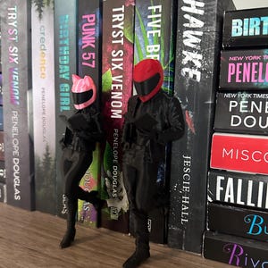 May include: Two figurines of people in black motorcycle gear and helmets, one pink and one red, stand in front of a bookshelf filled with books. The books have titles like "TRYST SIX VENOM" and "HAWKE".