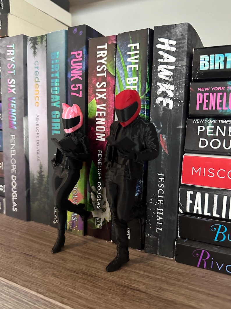 May include: Two figurines of people in black motorcycle gear and helmets, one pink and one red, stand in front of a bookshelf filled with books. The books have titles like "TRYST SIX VENOM" and "HAWKE".
