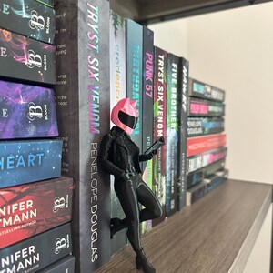 May include: A black figurine of a woman in a motorcycle helmet and outfit, leaning against a row of books. The books have titles like "TRYST SIX VENOM" and "FIVE BROTHERS". The figurine is on a wooden shelf, with other books visible.