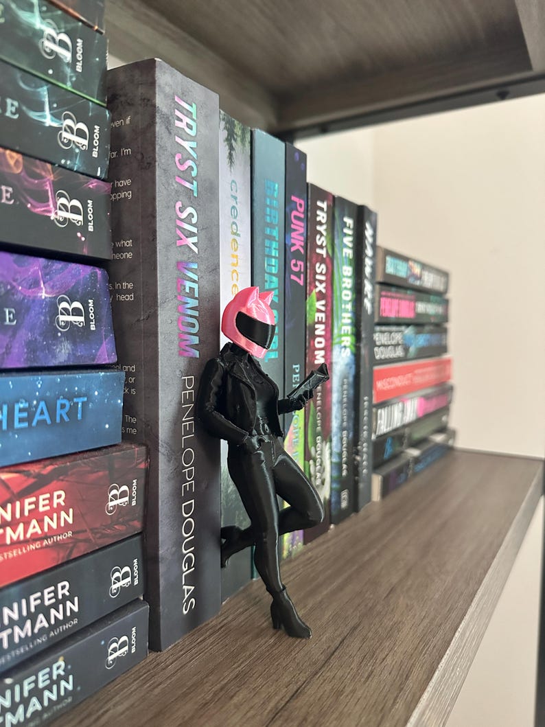 May include: A black figurine of a woman in a motorcycle helmet and outfit, leaning against a row of books. The books have titles like "TRYST SIX VENOM" and "FIVE BROTHERS". The figurine is on a wooden shelf, with other books visible.