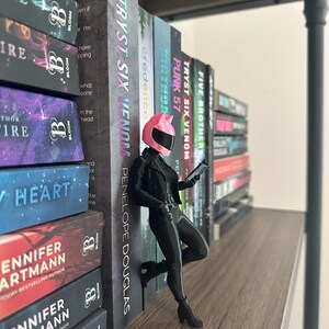 May include: A figurine of a person in a pink motorcycle helmet and black outfit, leaning against a shelf of books. The books have titles like "TRYST SIX VENOM" and "PUNK 57". The shelf is made of wood.