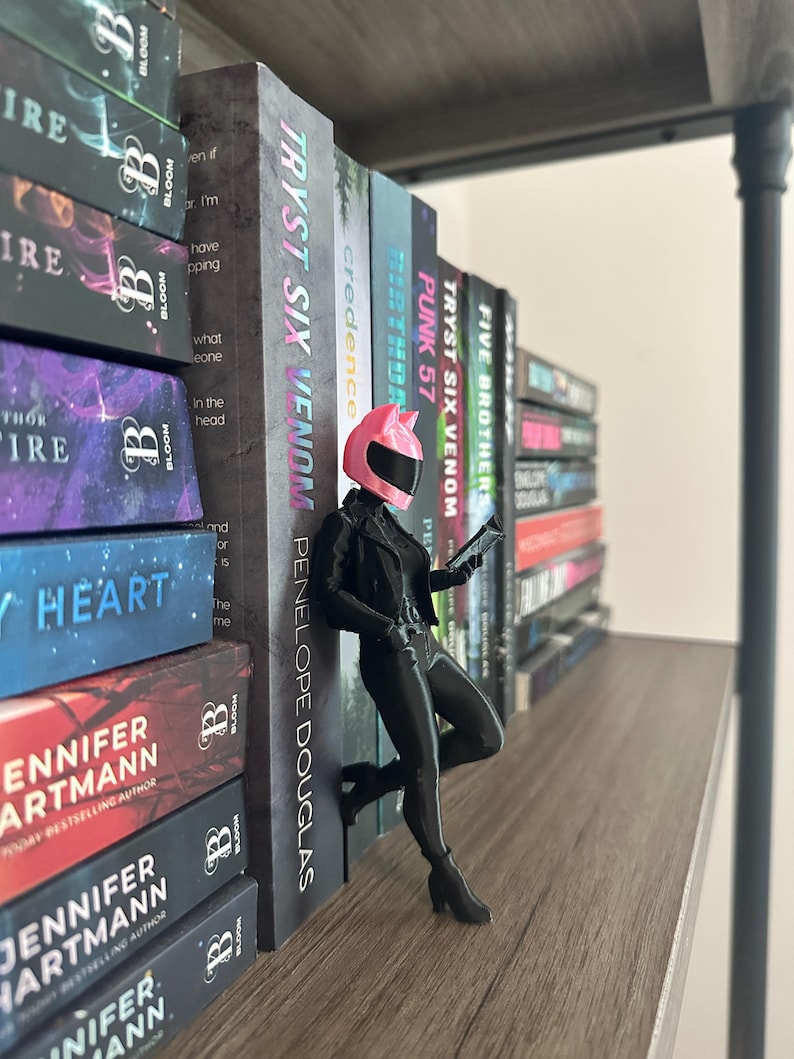 May include: A figurine of a person in a pink motorcycle helmet and black outfit, leaning against a shelf of books. The books have titles like "TRYST SIX VENOM" and "PUNK 57". The shelf is made of wood.