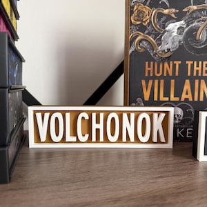 Puede incluir: Un letrero rectangular con el nombre "VOLCHONOK" en letras blancas en relieve sobre un fondo dorado. El letrero est&aacute; sobre una superficie de madera, con un libro titulado "HUNT THE VILLAIN" visible en el fondo.