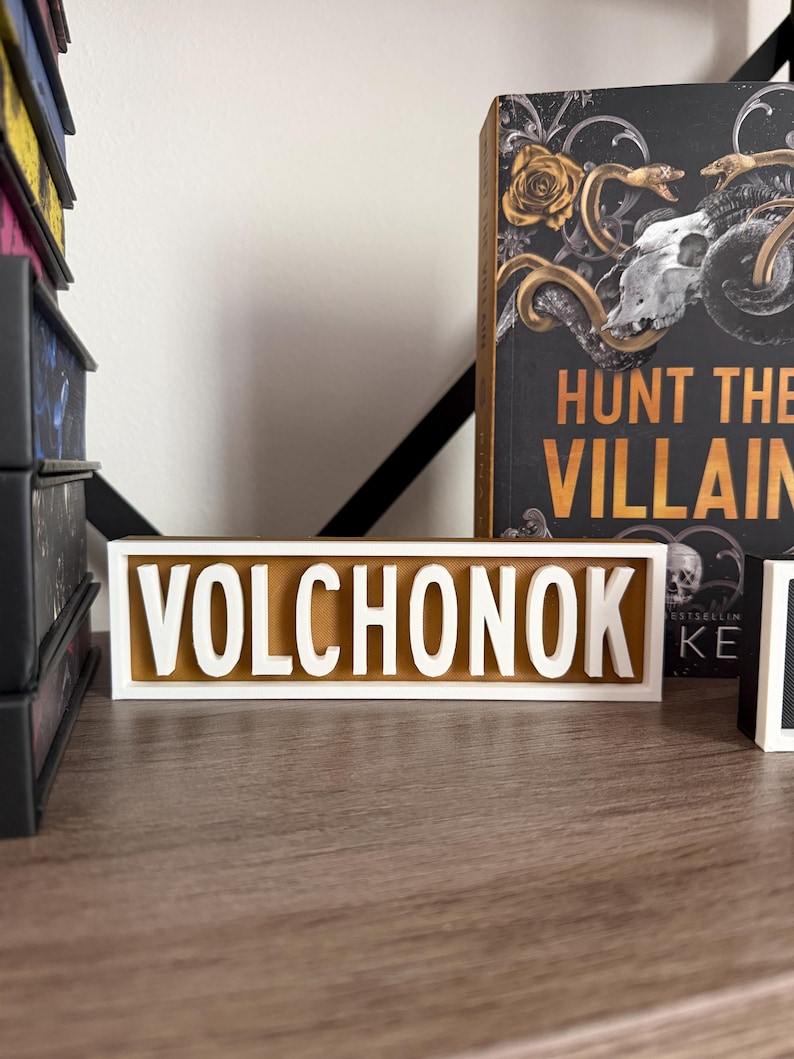 Puede incluir: Un letrero rectangular con el nombre "VOLCHONOK" en letras blancas en relieve sobre un fondo dorado. El letrero est&aacute; sobre una superficie de madera, con un libro titulado "HUNT THE VILLAIN" visible en el fondo.